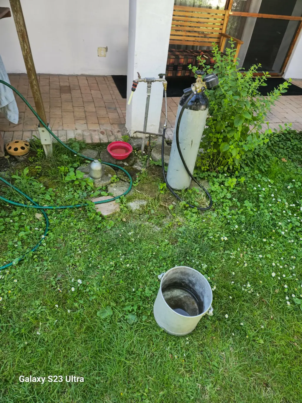 Nyomásszabályozóval felszerelt palack és csatlakozó tömlő egy családi ház kerti csapja mellett
