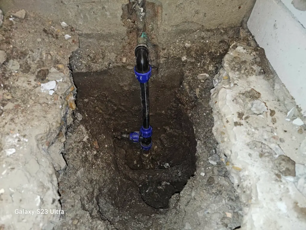 Betonburkolatban kibontott gödörben álló fekete vízvezetékcső kék szorítógyűrűs csatlakozókkal és egy kék fogantyús szerelvénnyel